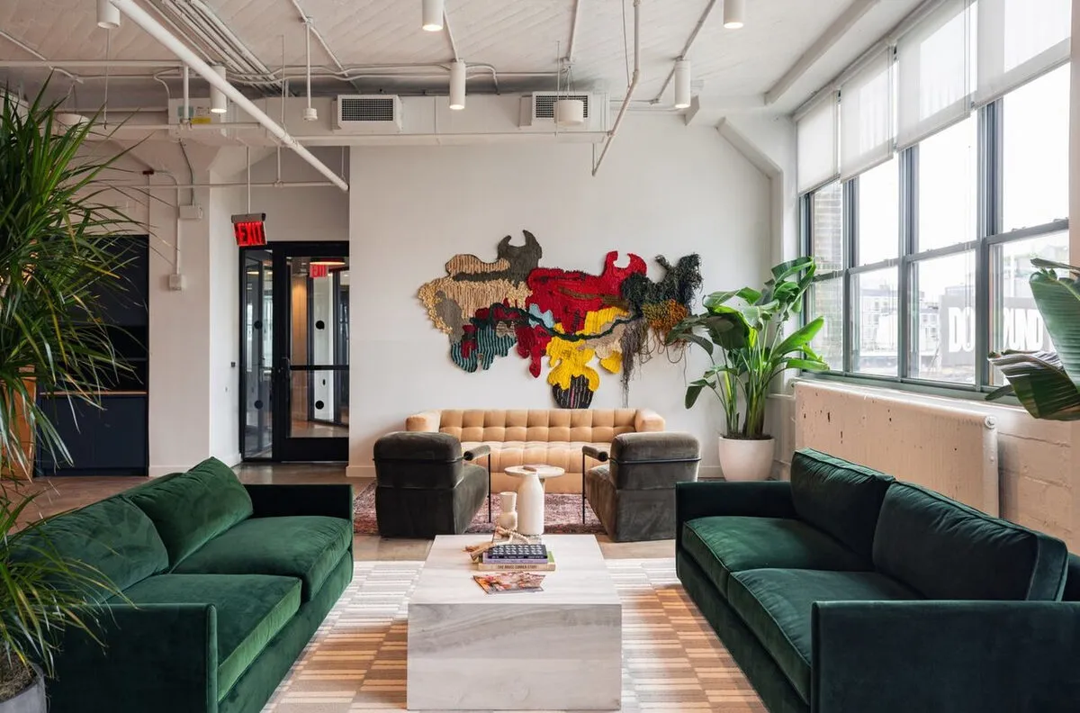 Loft office lounge with green velvet sofas and a large multicolored textile wall sculpture above a tufted sofa