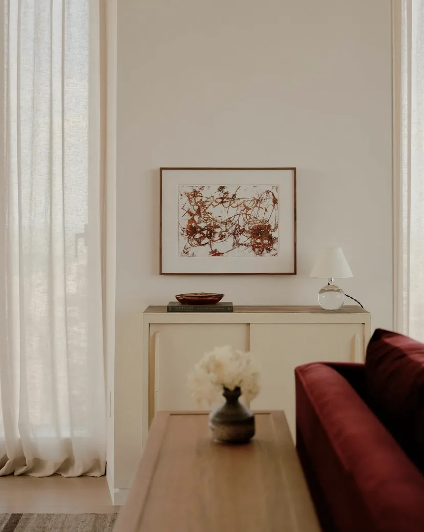 Small framed red and brown abstract drawing above a cream cabinet and lamp beside sheer curtains