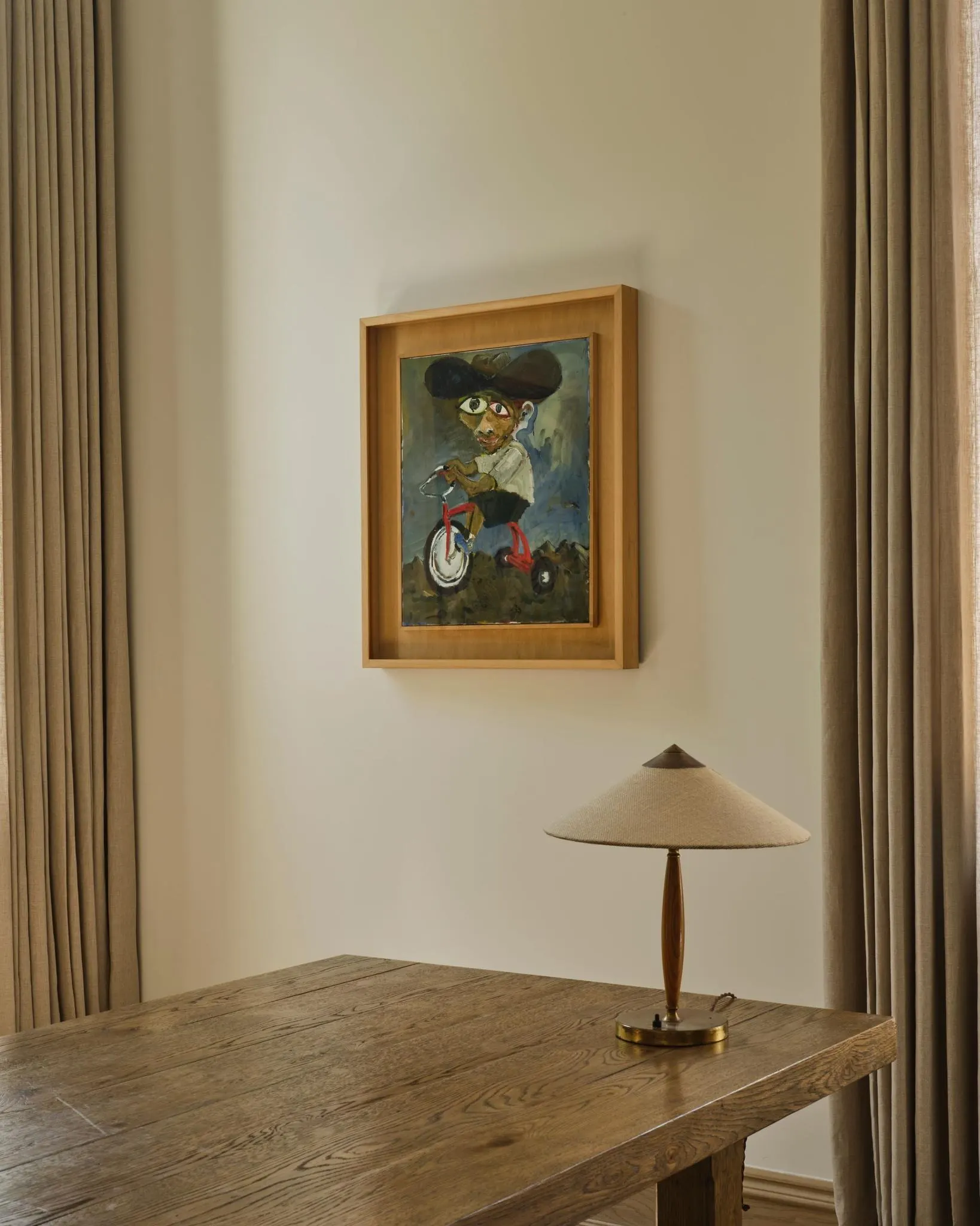 Framed painting of a figure in a wide hat riding a red tricycle above a wood table and lamp