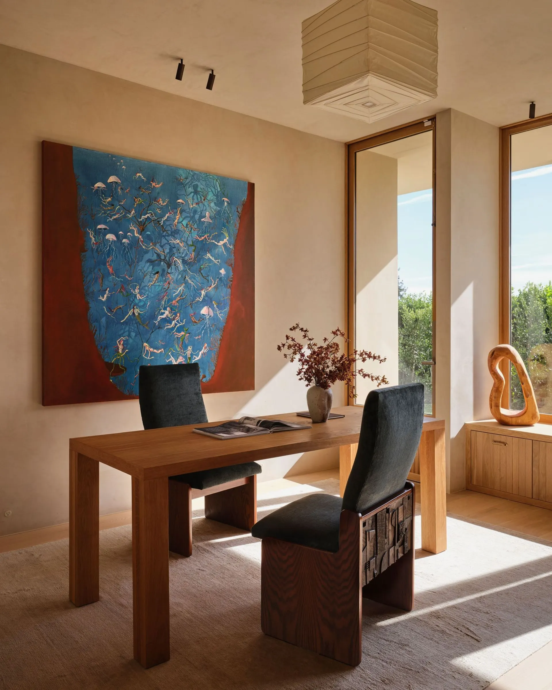 Large blue painting of small swimming figures framed in red above a wood desk in a sunny study