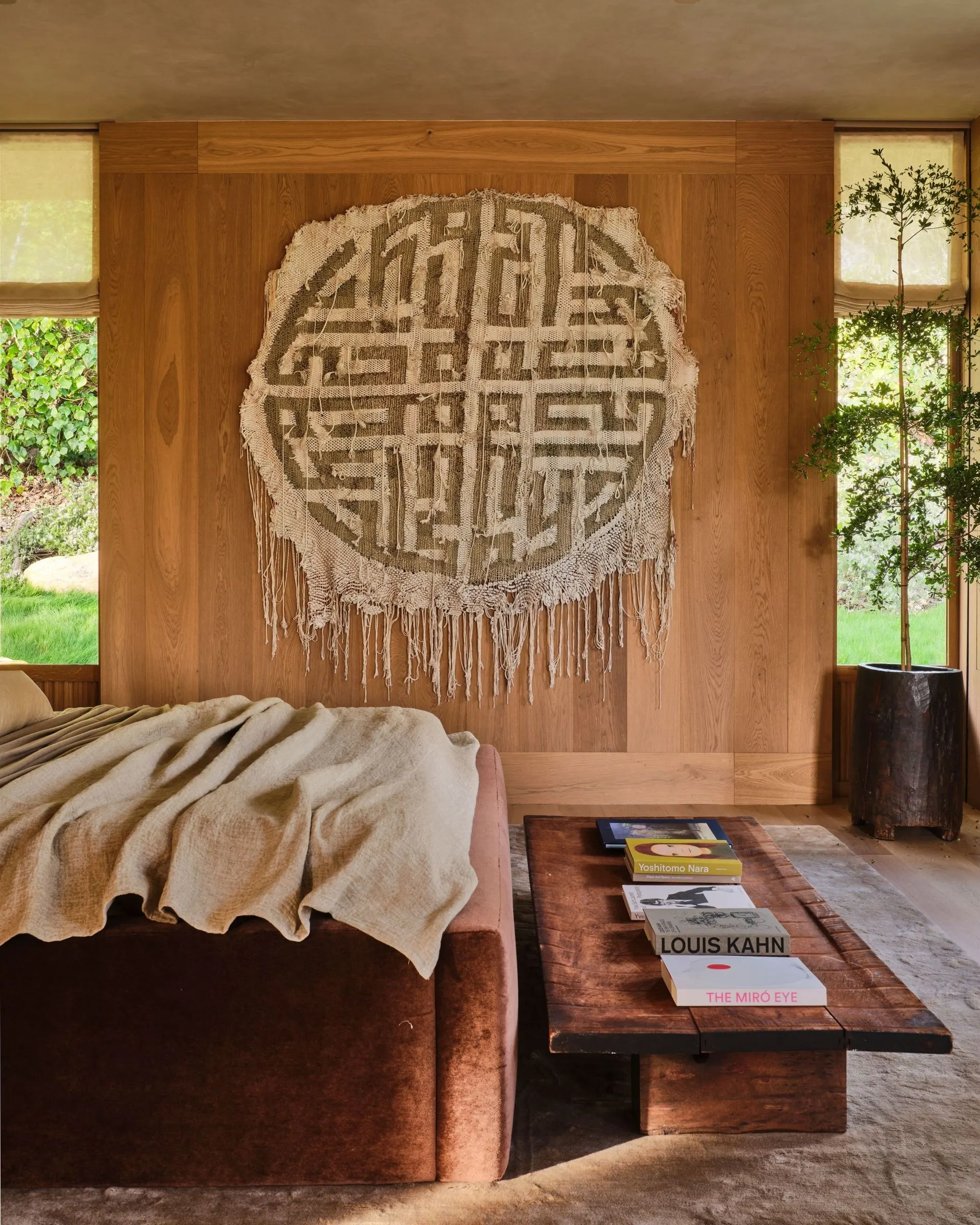 Large round fringed textile with a gray maze pattern on a wood wall behind a bed, artwork by Igshaan Adams