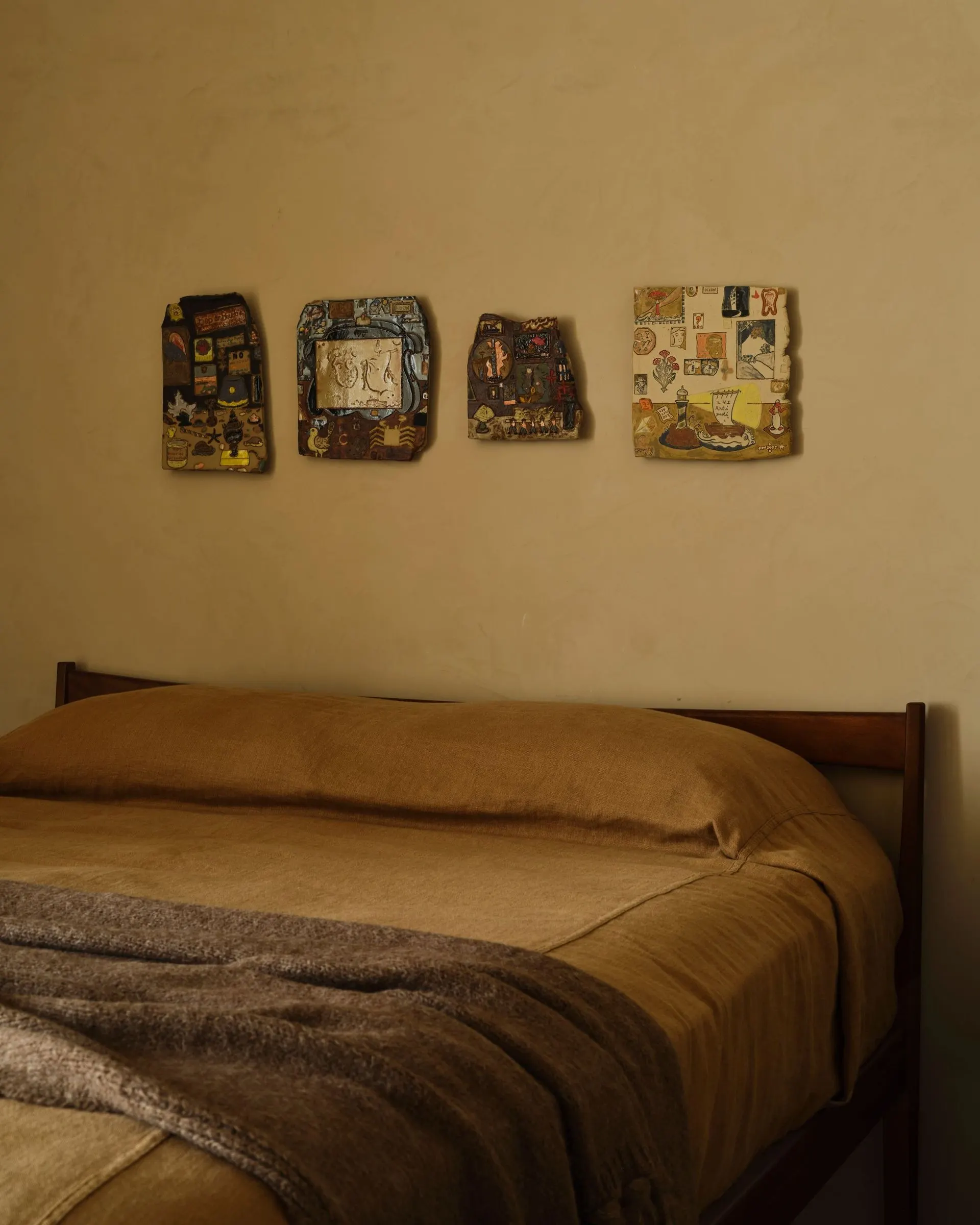 Four small irregular ceramic tiles with painted scenes hung above a bed with brown linens, artwork by Kevin McNamee-Tweed
