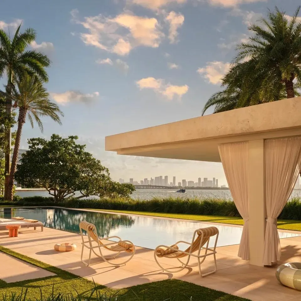 Waterfront pool terrace with lounge chairs and a curtained pavilion, palm trees and a city skyline beyond