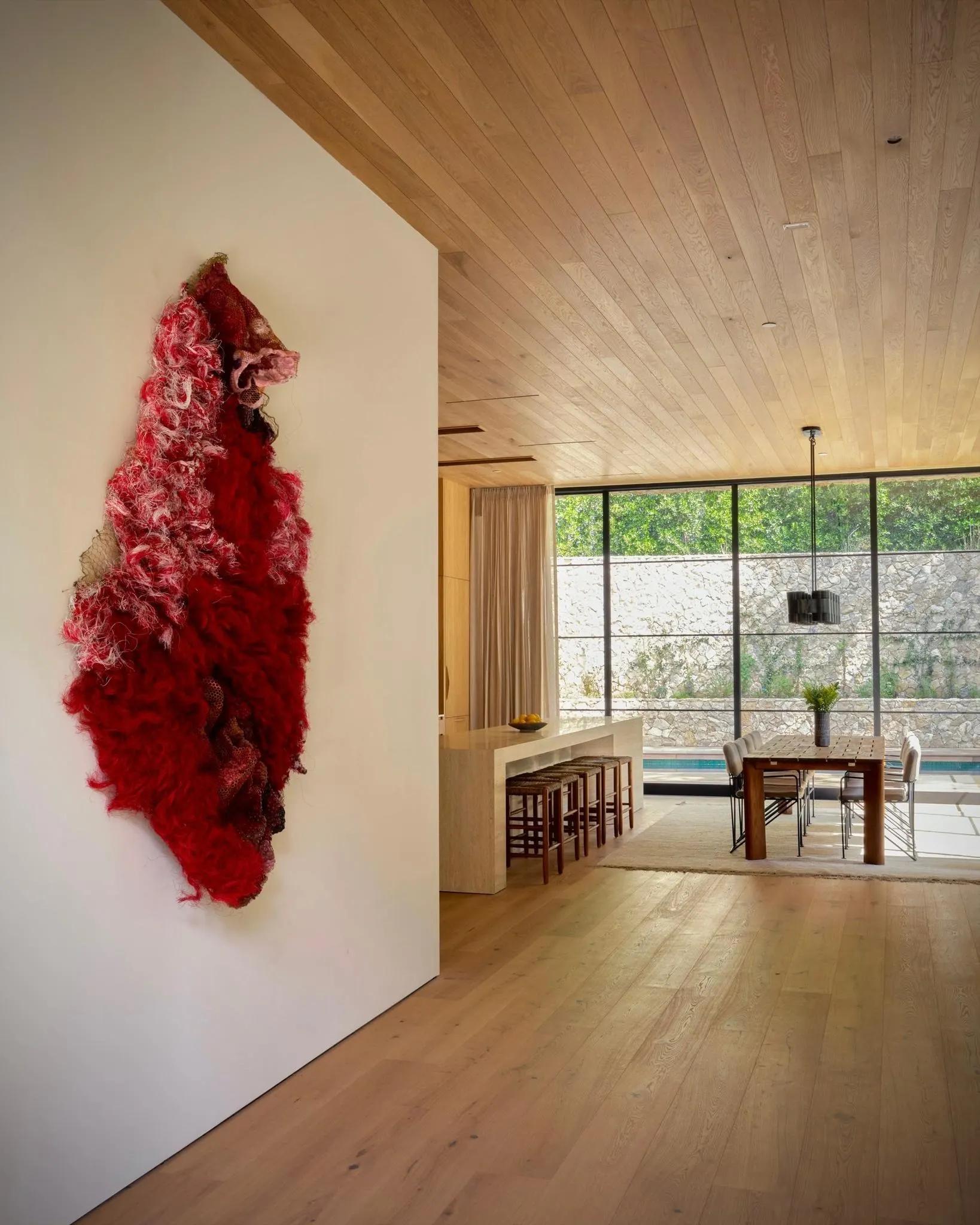 Red shaggy textile wall sculpture on a white wall beside an open kitchen and dining area, artwork by Julio Rizhi