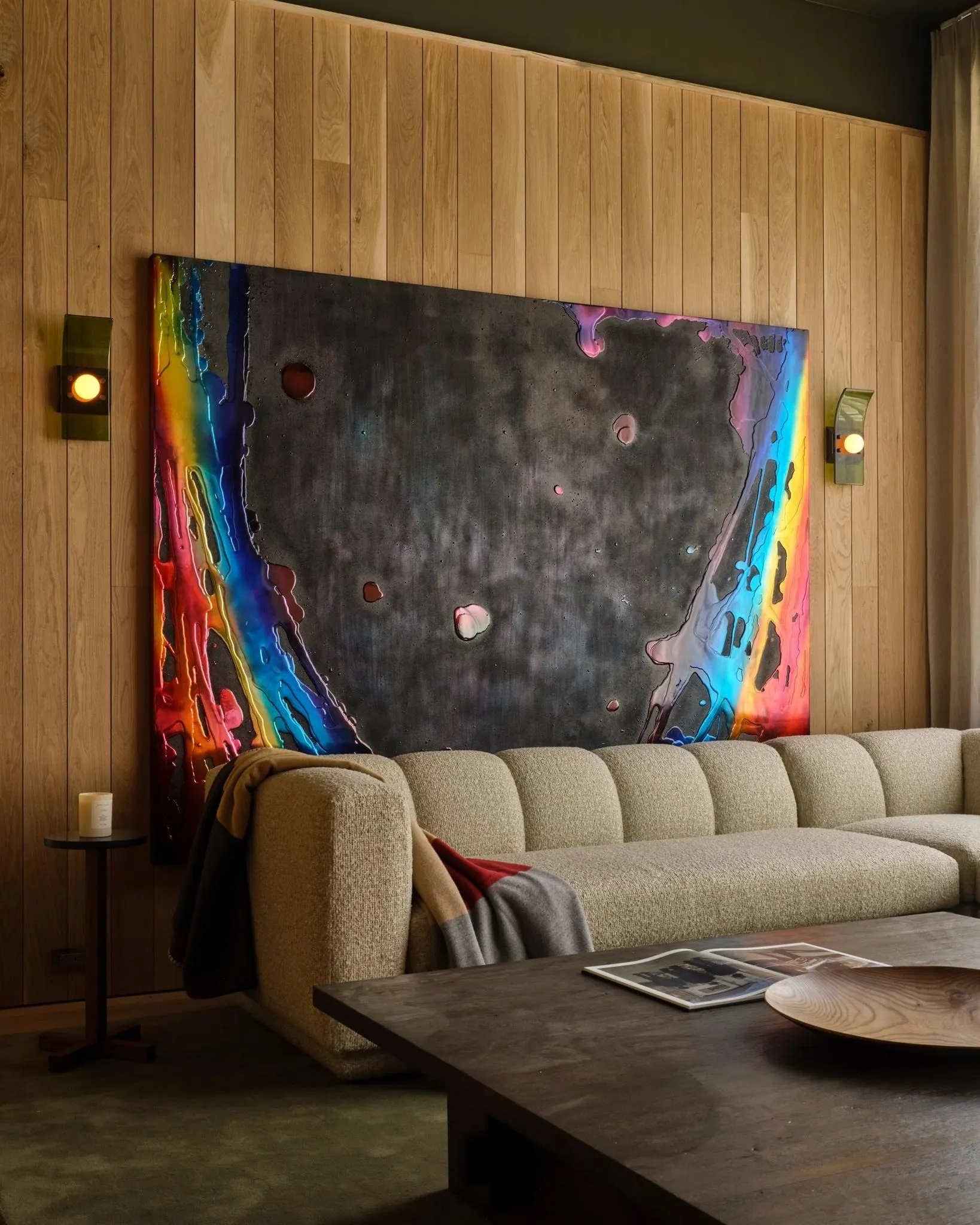 Dark abstract painting edged with rainbow drips above a cream sofa in a wood-paneled room, artwork by John Monn
