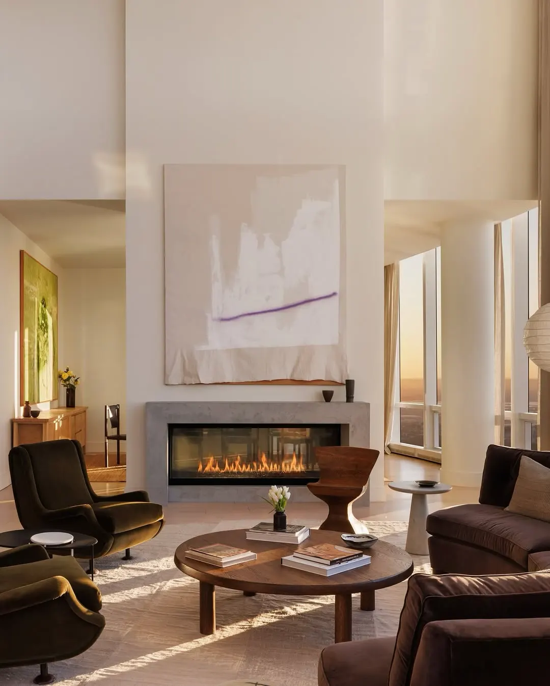 Sunlit double-height living room with a pale abstract canvas above a fireplace, velvet lounge chairs and skyline views