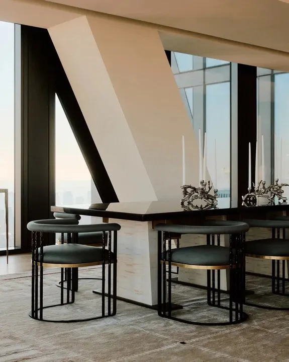 Dining table with sculptural silver candelabras and dark green chairs beside a slanted column and tall windows