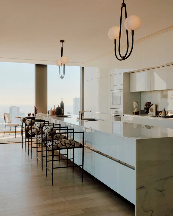 Kitchen with a long marble island, patterned bar stools and globe pendants beside floor-to-ceiling city windows