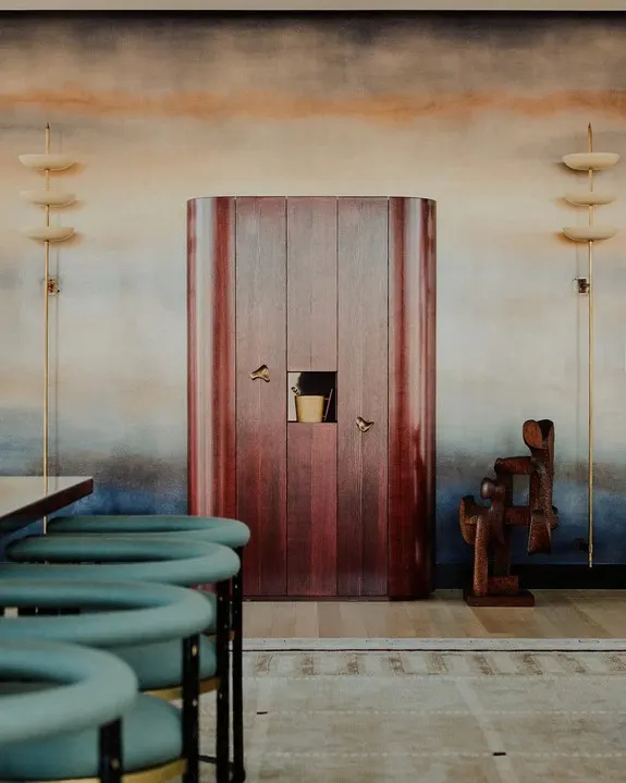 Rounded red wood cabinet and a carved wooden sculpture against an ombre painted mural wall with brass sconces