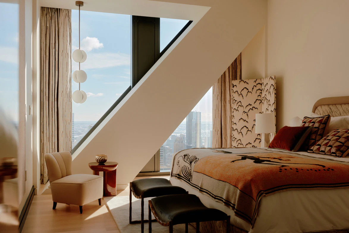 Bedroom with a slanted window wall, globe pendant, patterned orange blanket and a cream armchair overlooking Manhattan