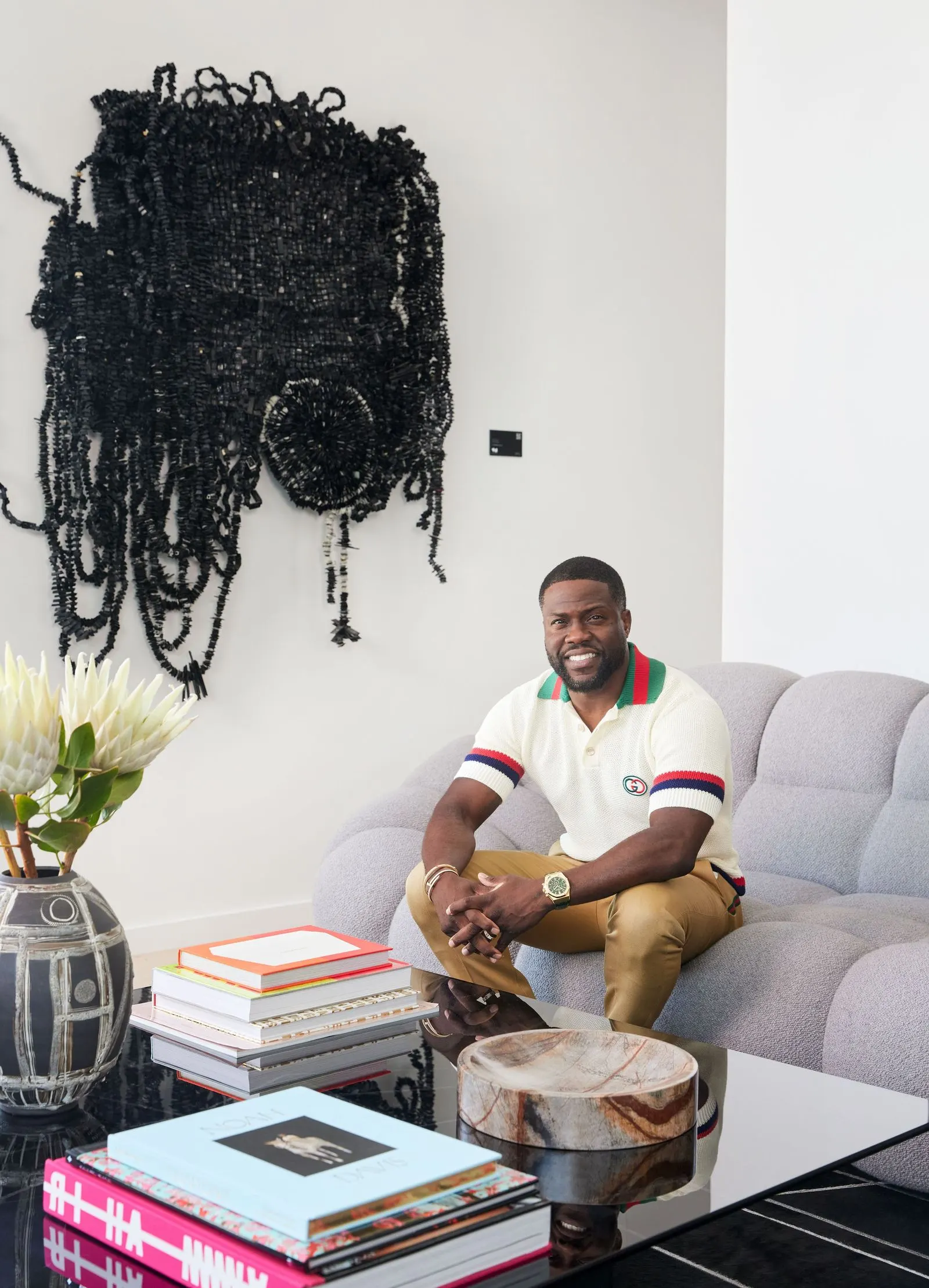 Kevin Hart seated on a gray sofa below a large black beaded wall sculpture in his West Hollywood office