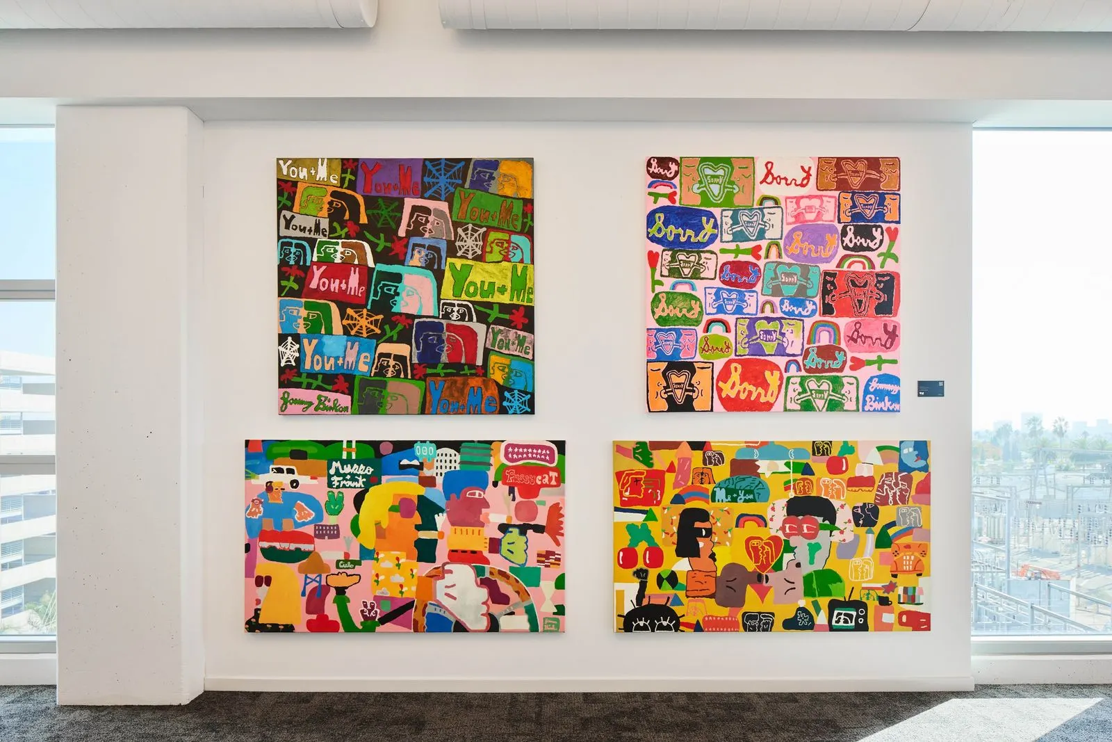 Four colorful pop art paintings with cartoon figures and bold lettering hung on a white office wall