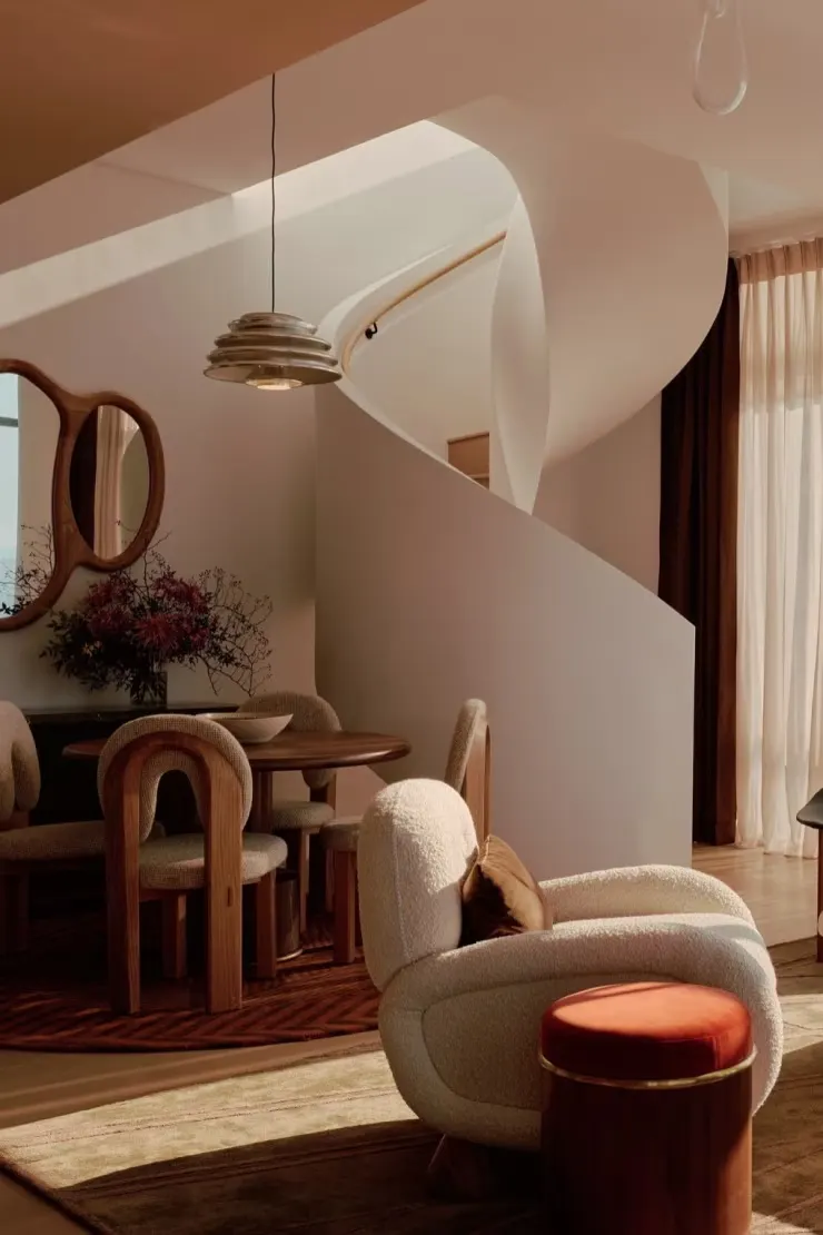 Dining area and curving white staircase with a sculptural wall mirror and a boucle armchair