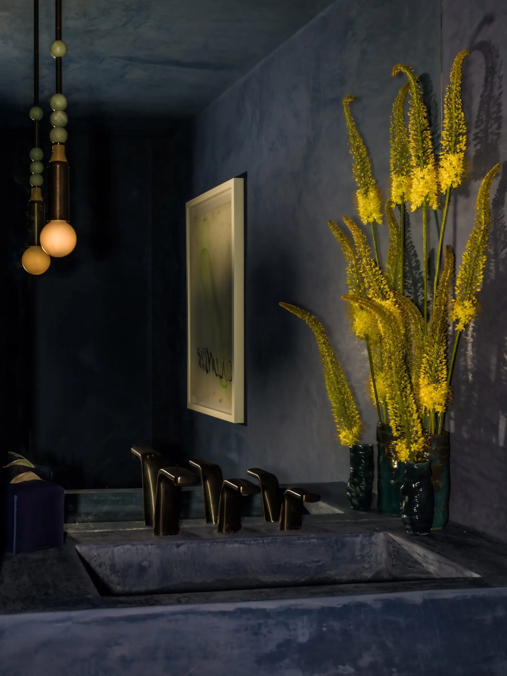 Dark blue-gray powder room with a stone trough sink, brass faucets, yellow flowers and a pale framed abstract