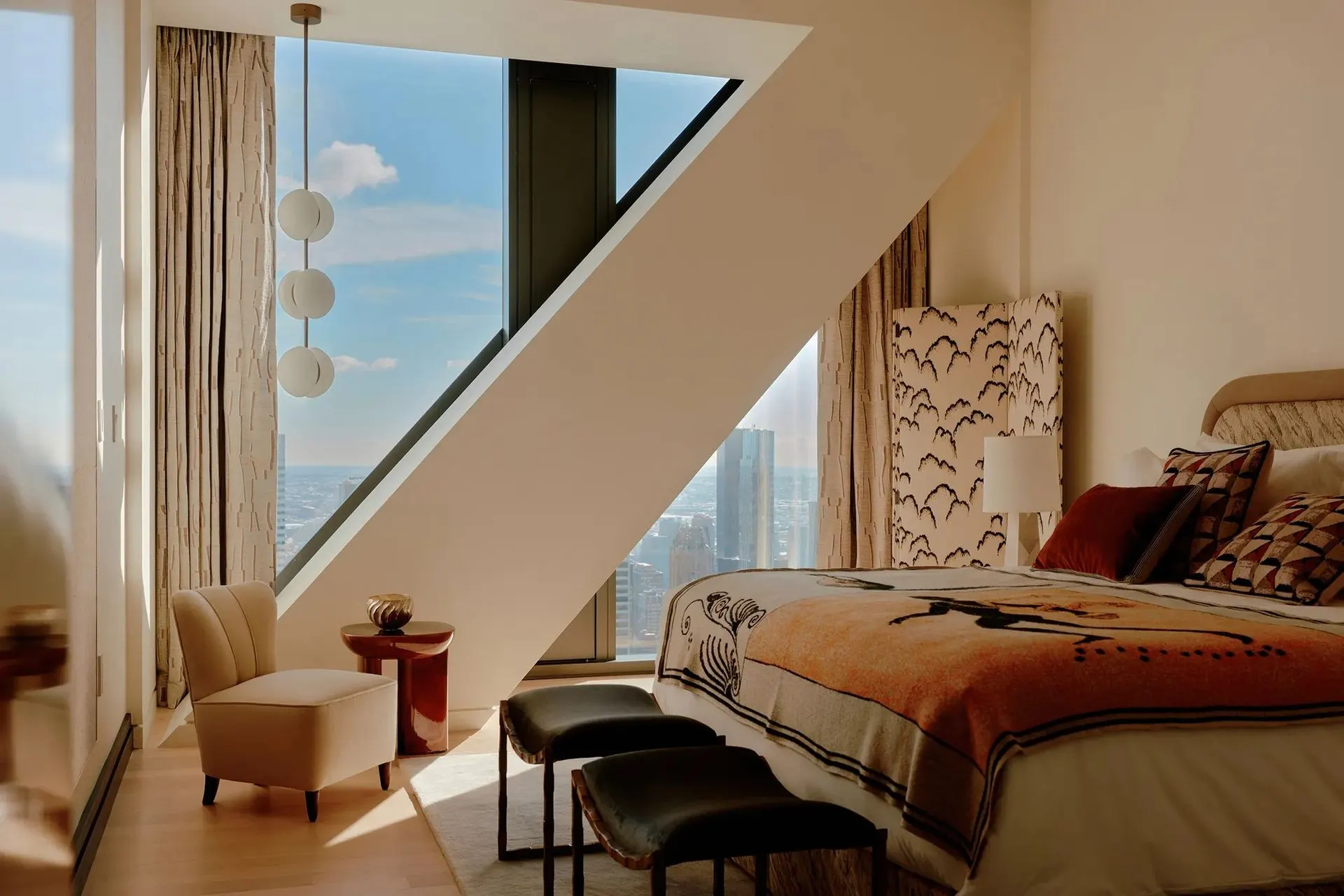 Bedroom with a slanted window wall, globe pendant, orange patterned blanket and cream armchair above Manhattan