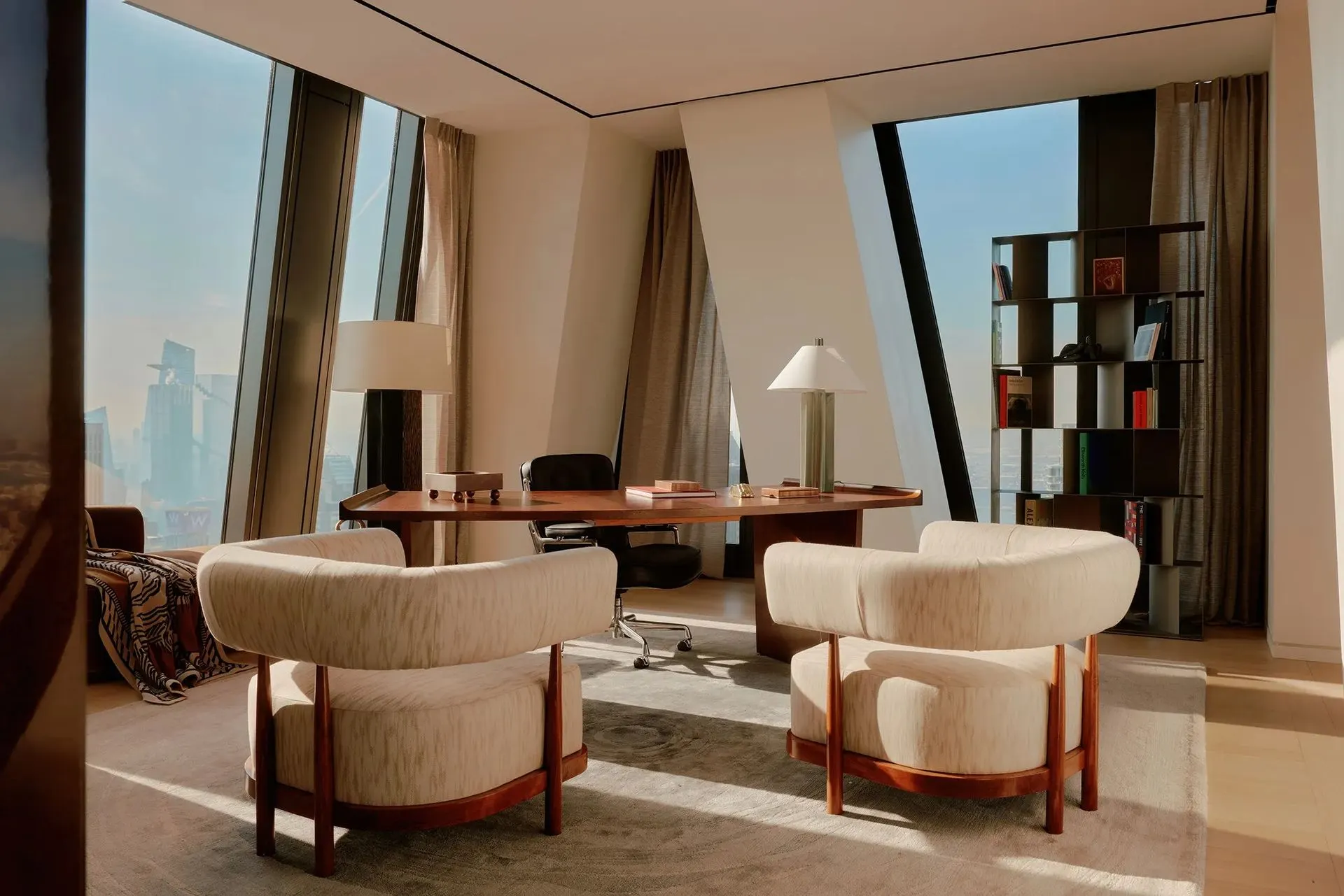 Home office with two cream boucle chairs before a walnut desk, bookshelves and slanted windows above the city