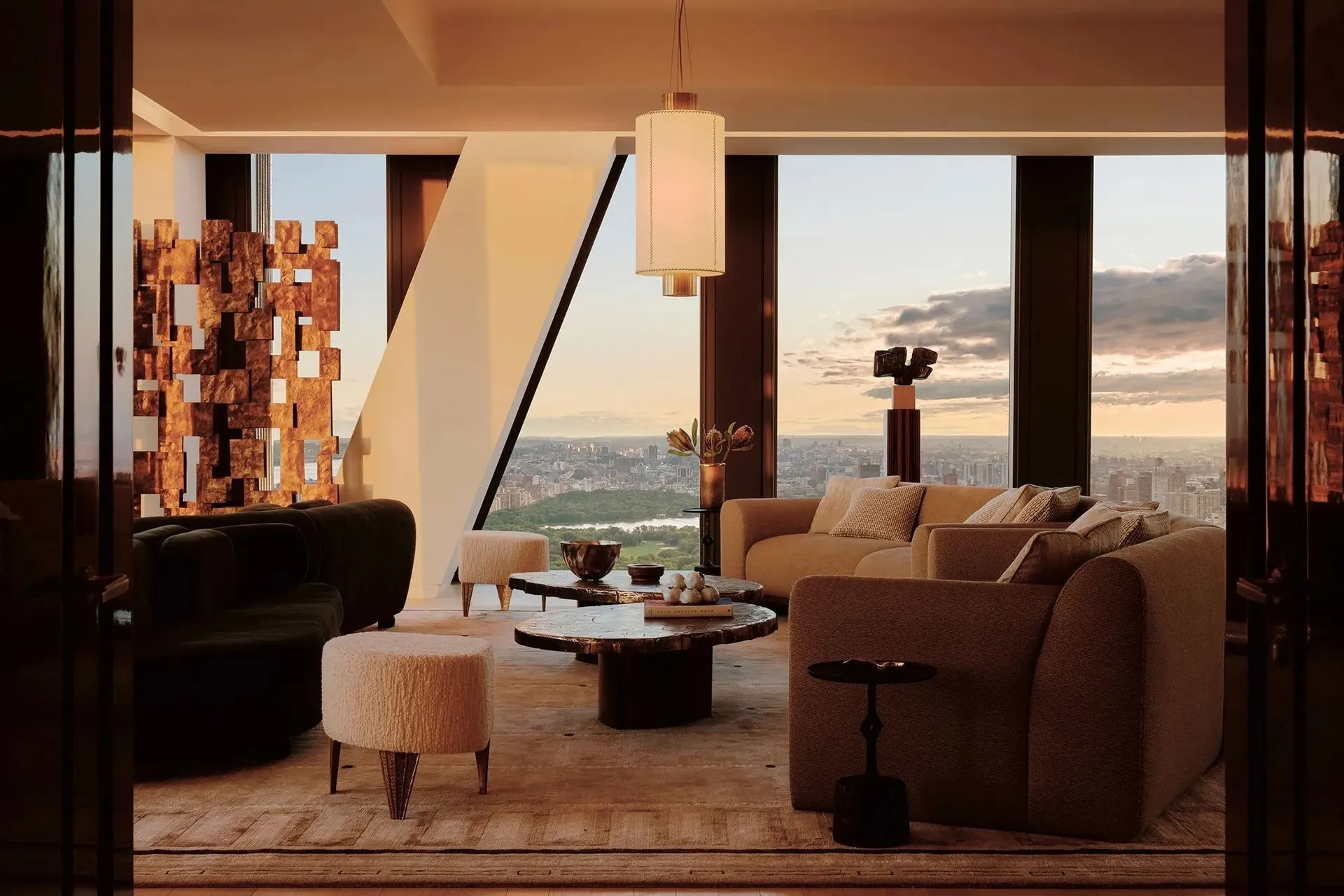 Living room with curved sofas, a bronze relief sculpture and floor-to-ceiling windows overlooking the city at dusk