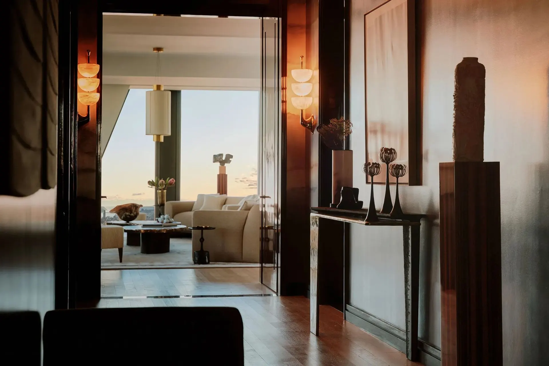 Dim entry hall with glowing alabaster sconces and a sculpture-topped console leading to a living room at sunset