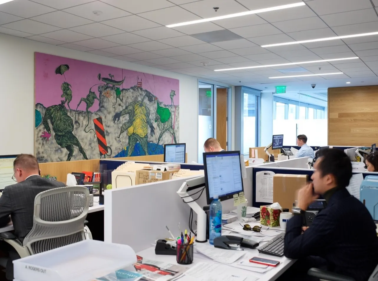 Open-plan office desks beneath a large pink mural-style painting of green cartoon creatures