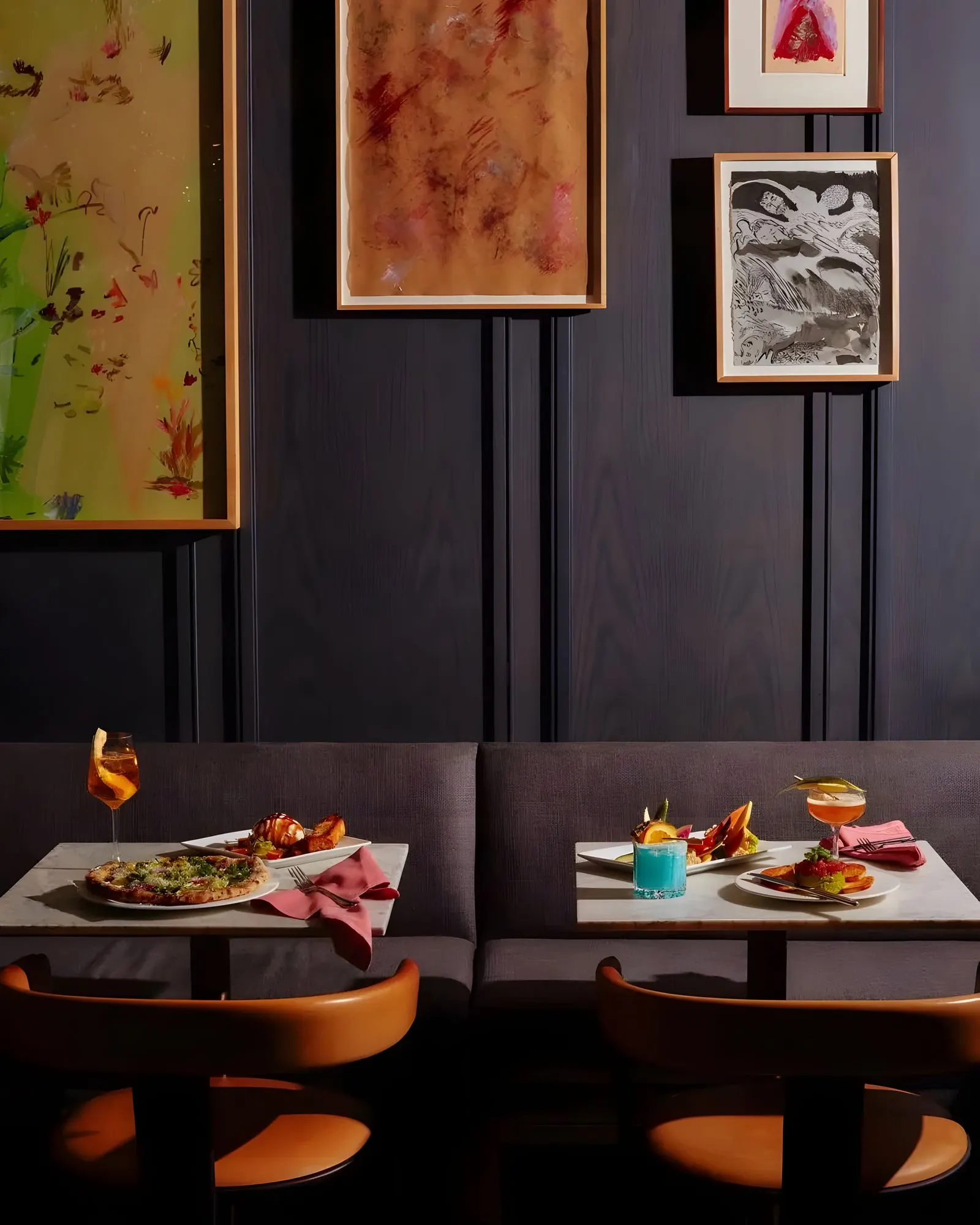 Restaurant banquette set with food and cocktails beneath framed abstract works on paper on dark paneled walls