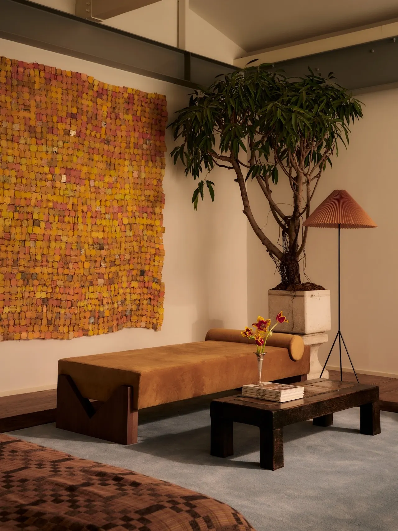 Bedroom corner with a large orange and yellow woven textile artwork above a suede daybed and potted tree