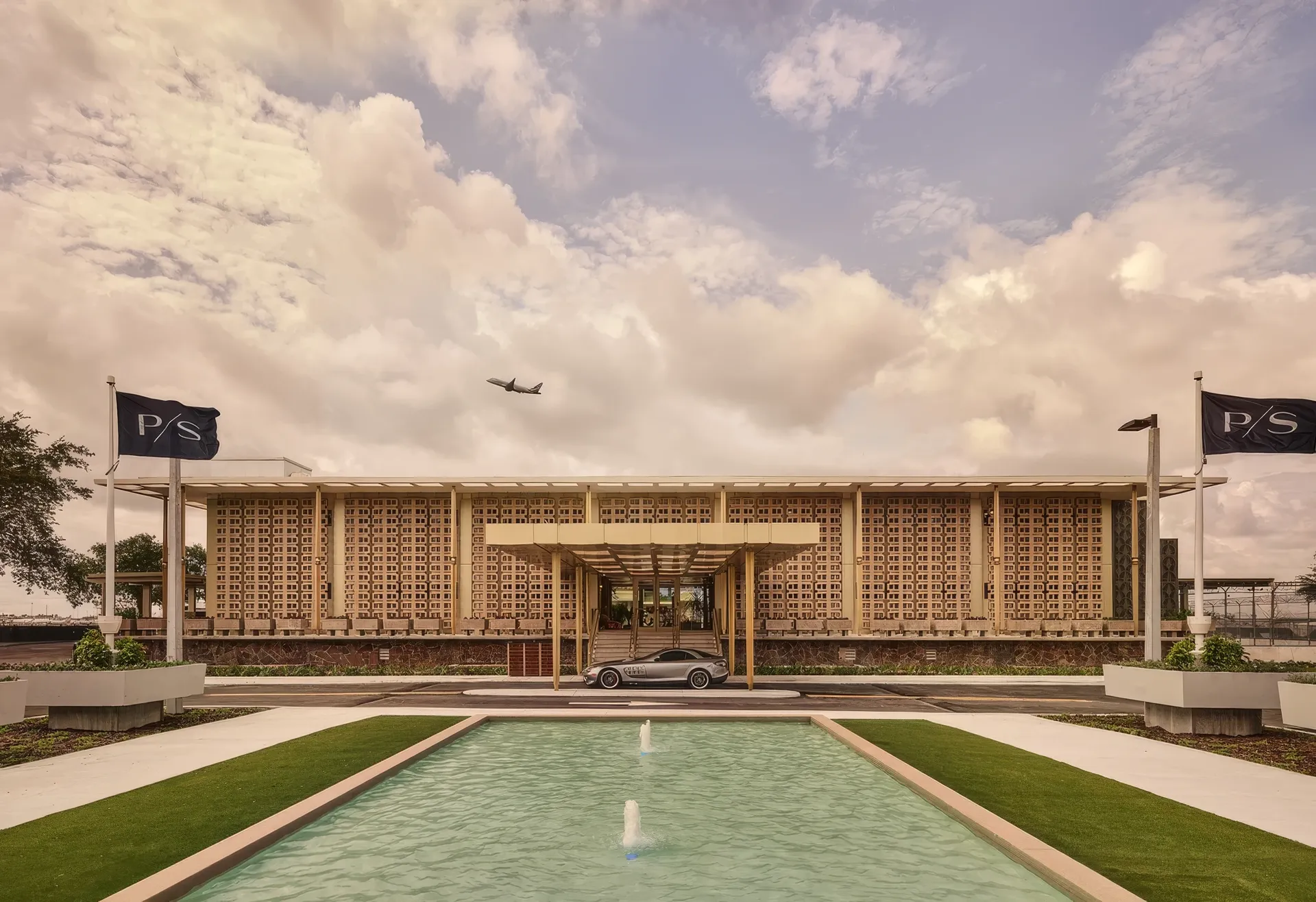 Mid-century private airport terminal in Miami with a reflecting pool, flags and a plane overhead