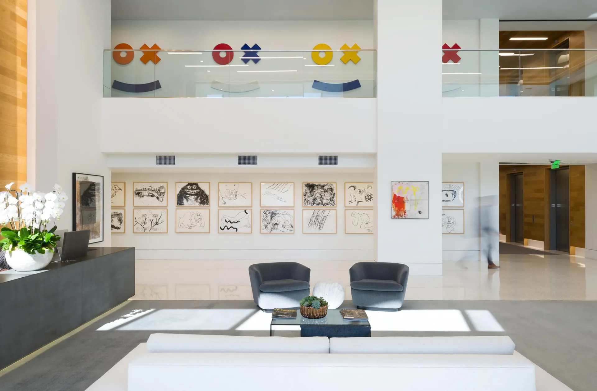 Double-height corporate lobby with a grid of framed drawings and colorful wall sculptures on the mezzanine