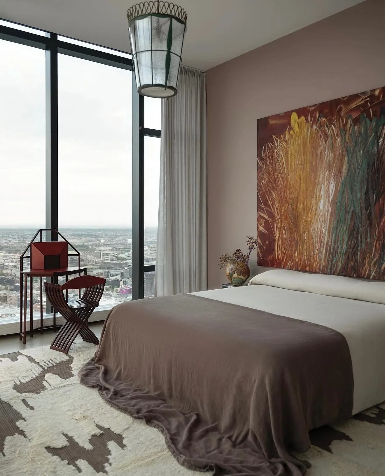 Bedroom with a large red, gold and green abstract painting above the bed beside floor-to-ceiling city windows