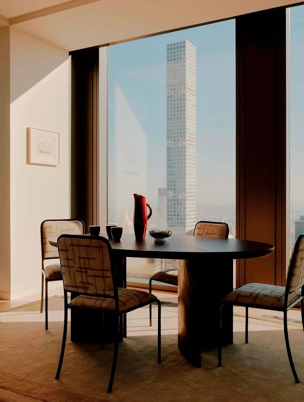Round dark dining table with patterned chairs and a red pitcher beside tall windows framing a Manhattan skyscraper