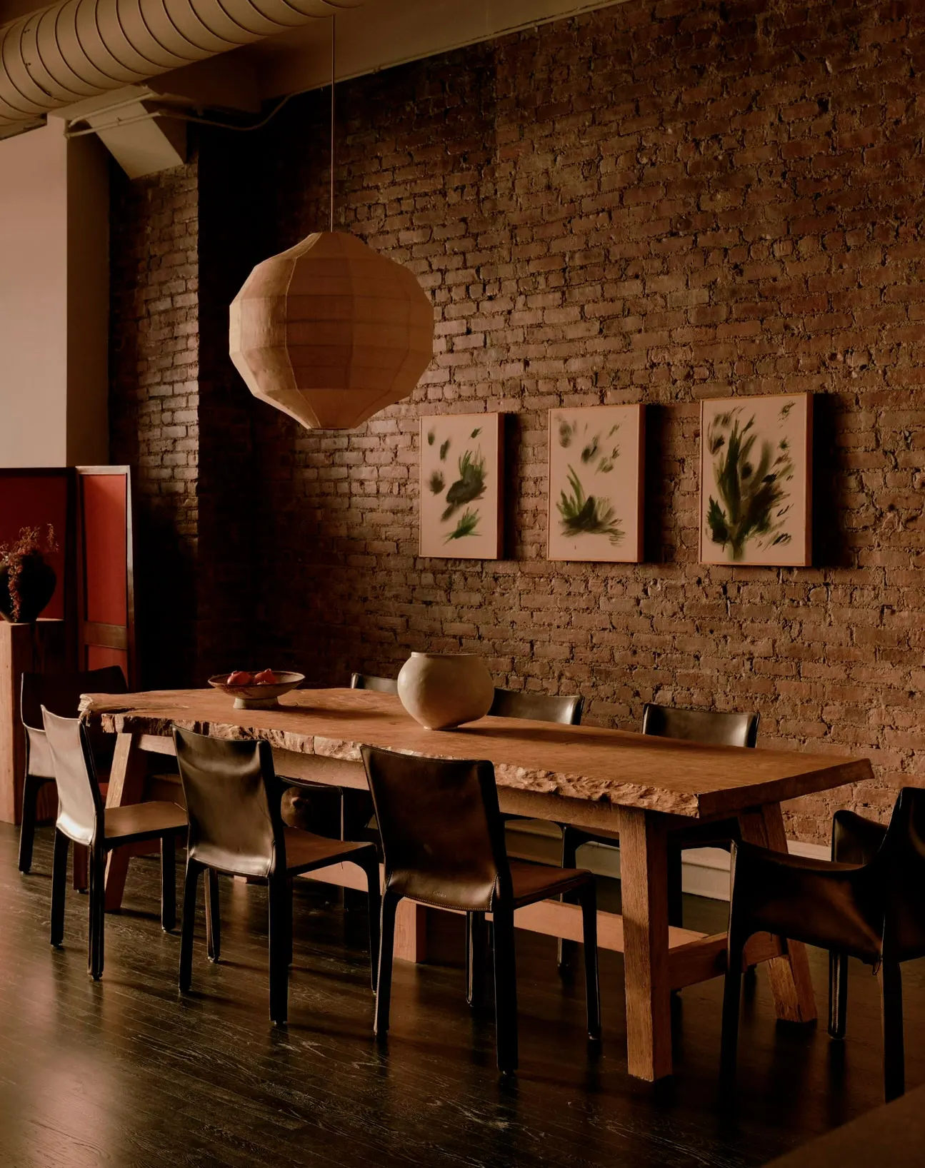 Loft dining room with three framed green botanical paintings on a brick wall above a long live-edge table