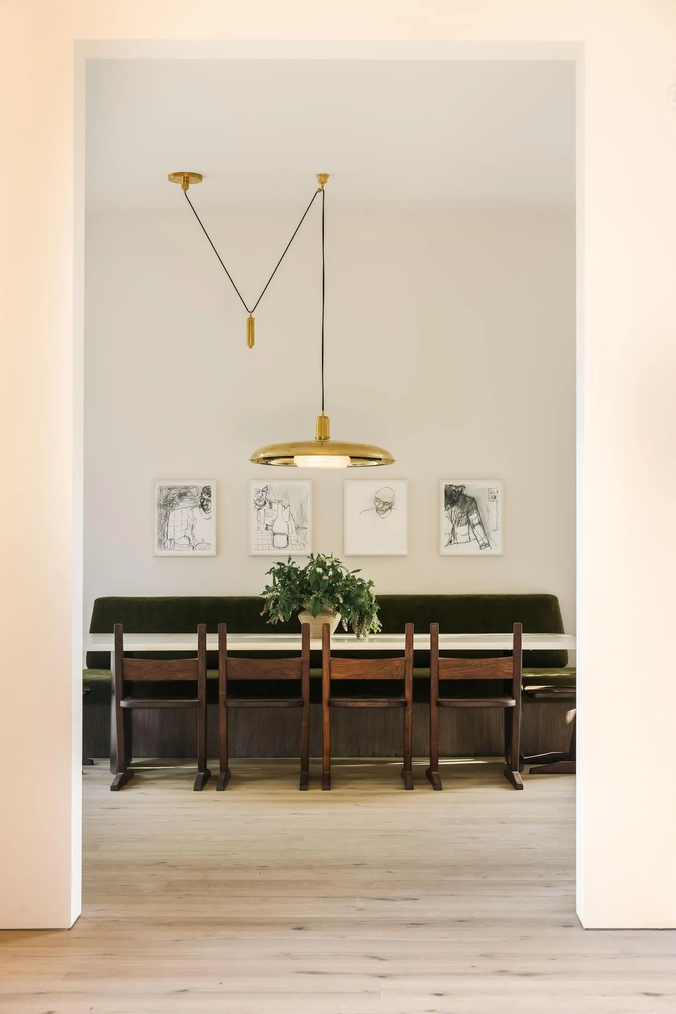 Dining nook with four framed black and white drawings above a green banquette, wooden chairs and brass pendant