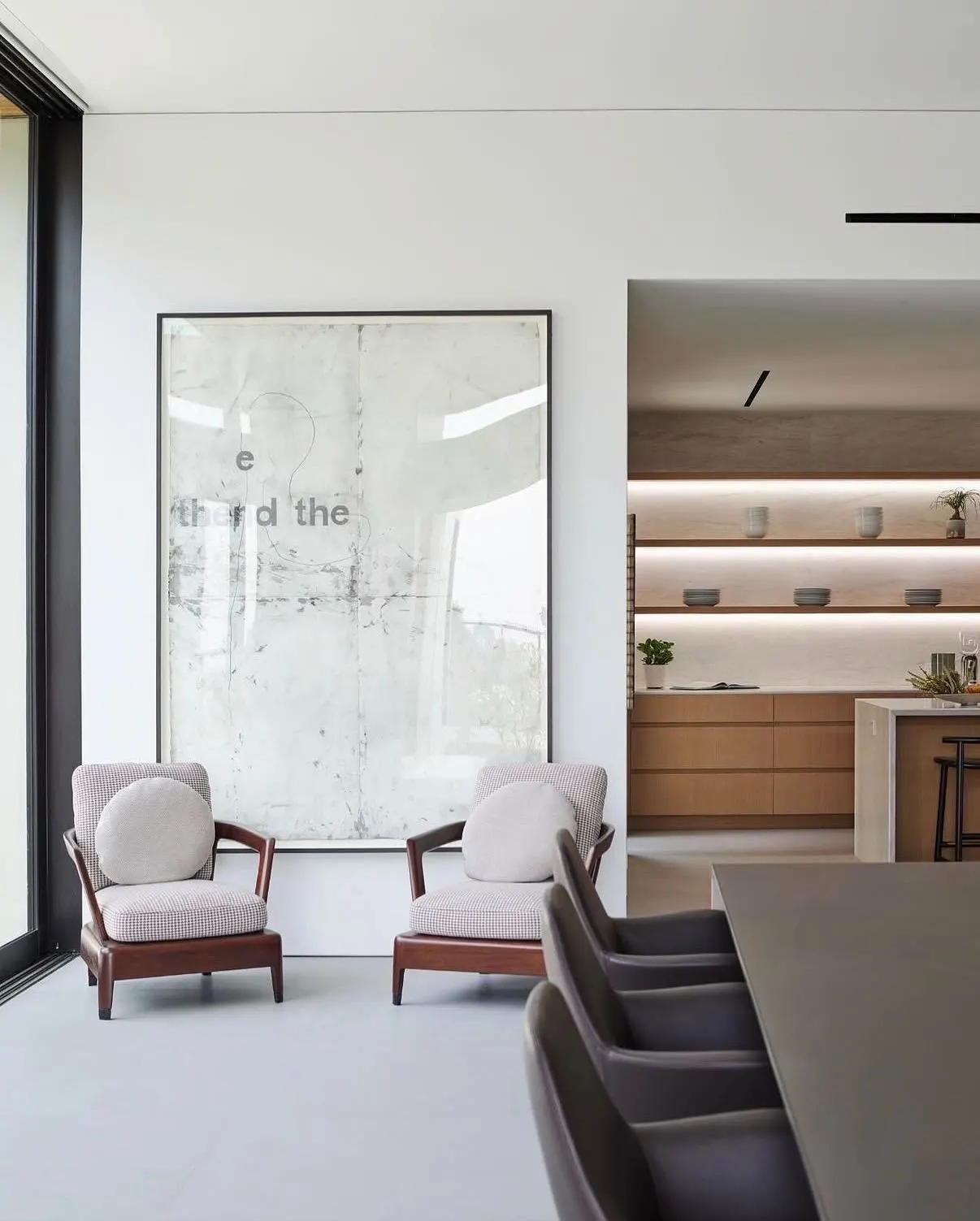Large pale text-based artwork behind two pink upholstered armchairs beside a dining table and open kitchen