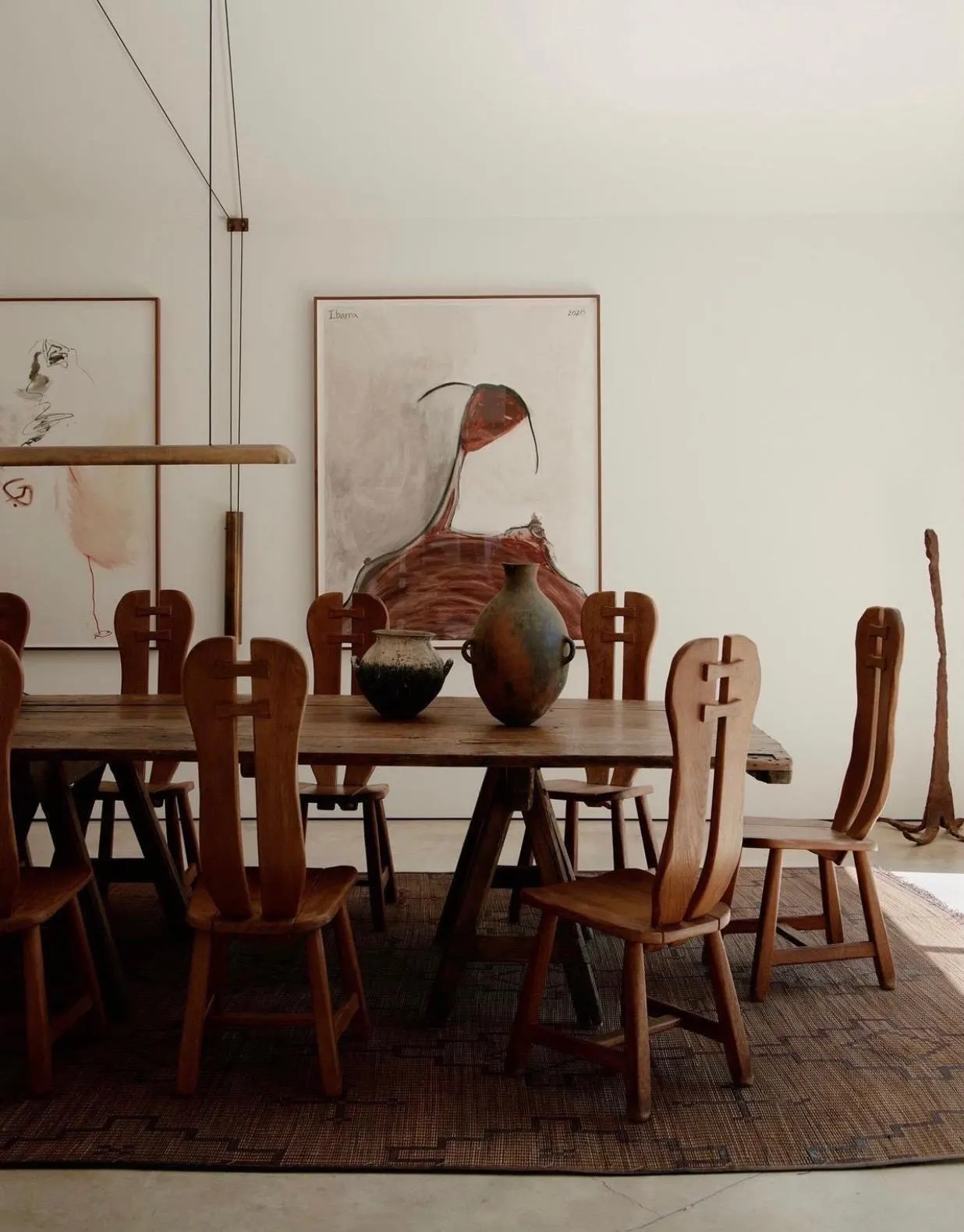 Dining room with two framed figurative drawings in red and black above a rustic wooden table and carved chairs
