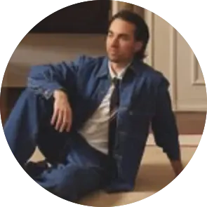 Interior designer Jake Arnold seated on the floor in a blue denim jacket and tie