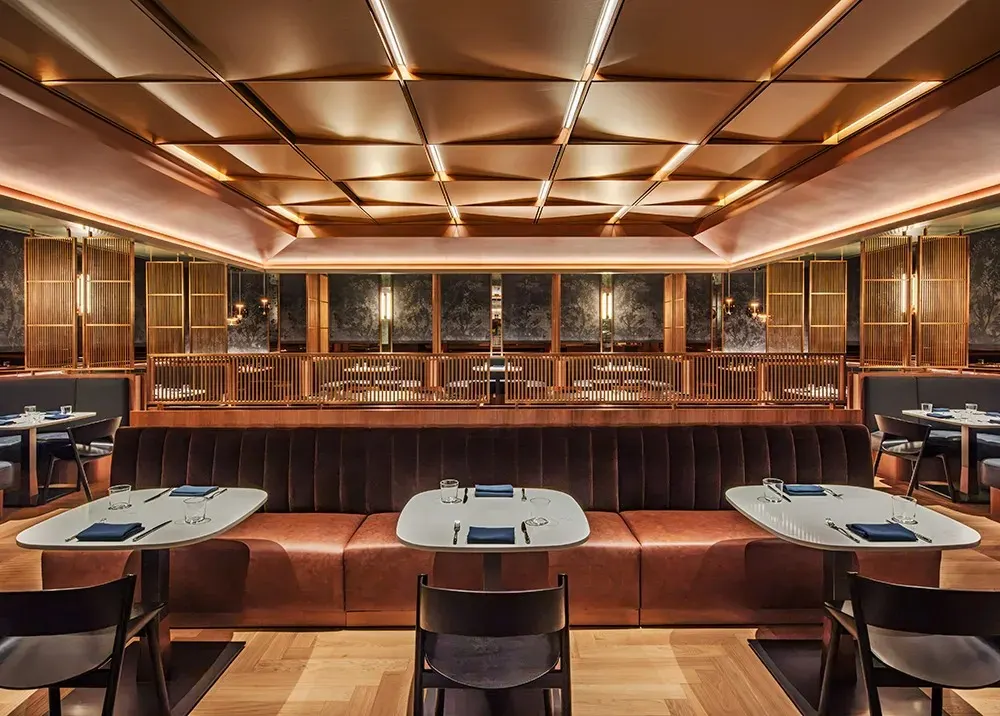 Restaurant dining room with a curved leather banquette, white tables and a glowing coffered copper ceiling