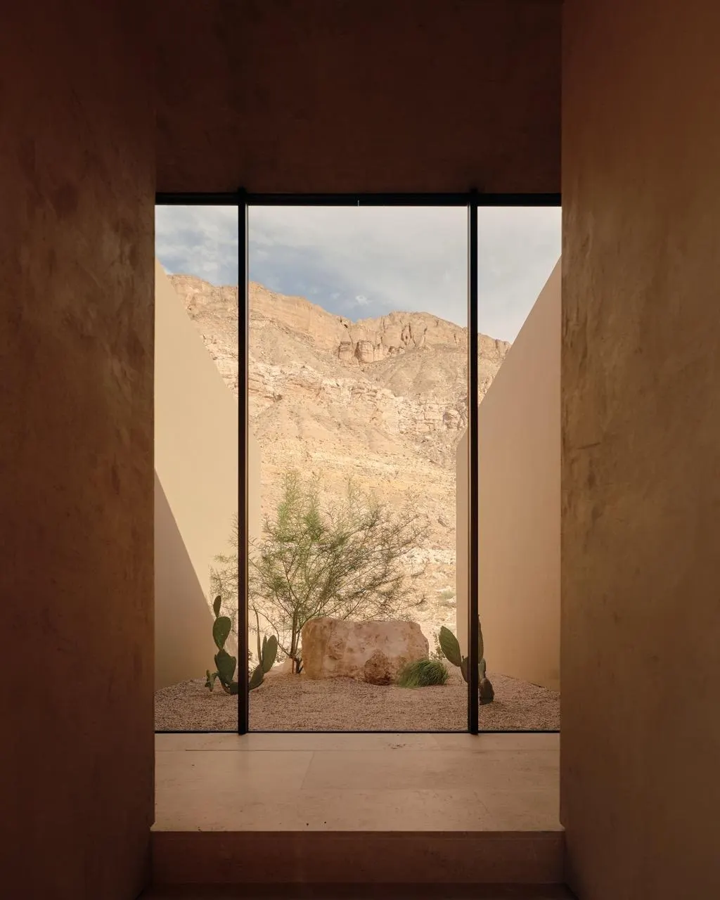 Floor-to-ceiling window framing a desert courtyard with cactus, a boulder and red rock cliffs beyond
