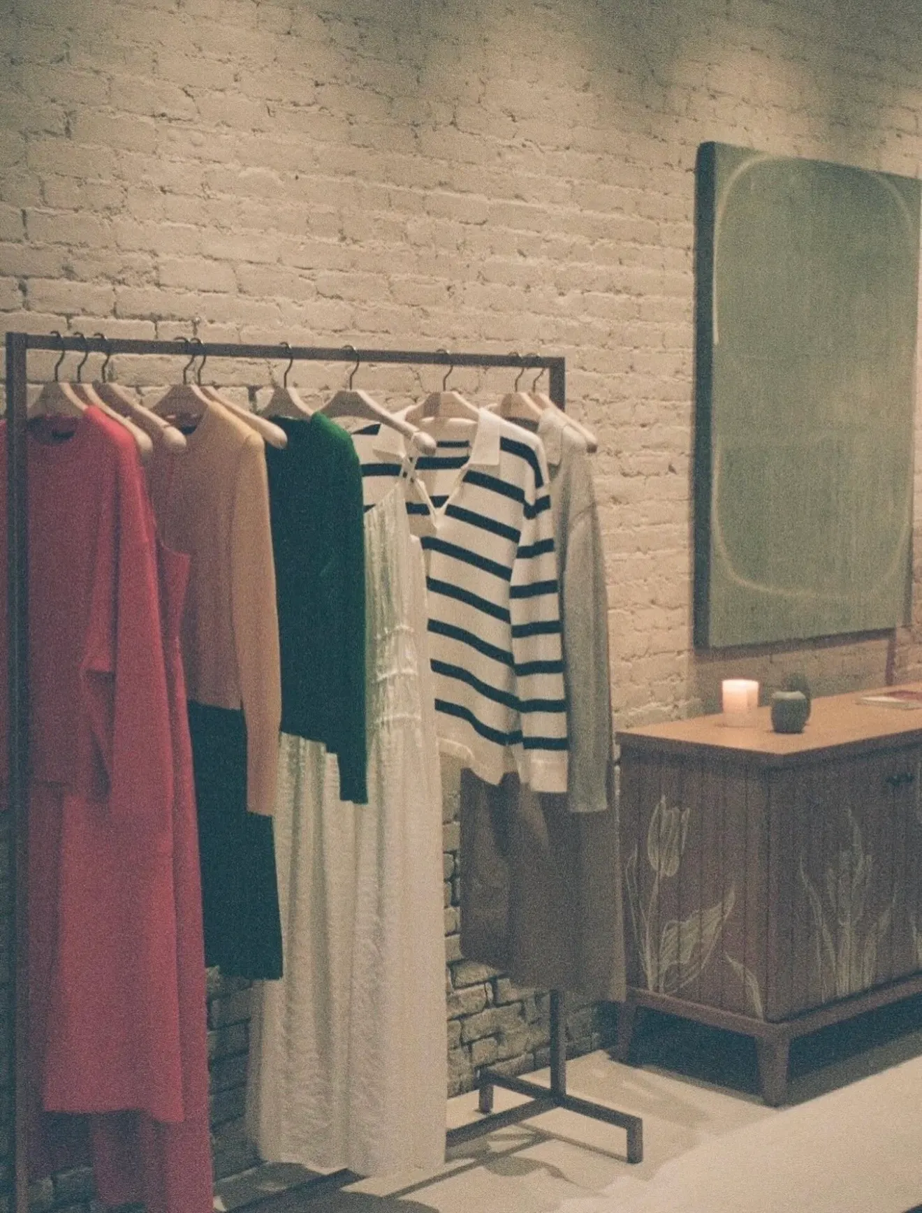 Clothing rack of knitwear and dresses beside a large green abstract painting on a white brick wall in a retail boutique